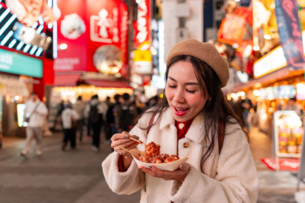 Mujer joven disfrutando de comida callejera en una animada calle comercial de Japón.