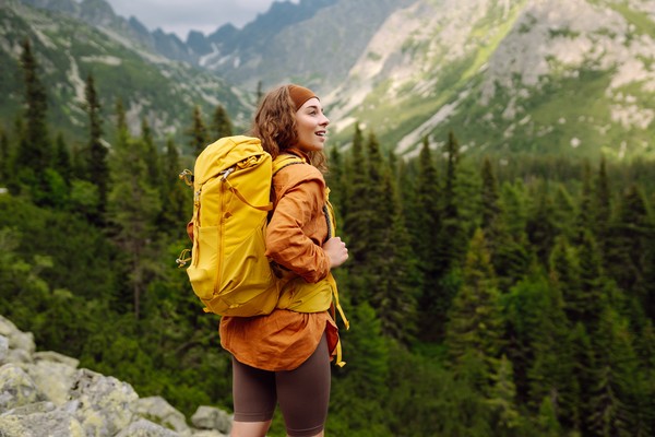 Una viajera sonriente con una mochila de senderismo amarilla disfrutando de un paisaje de montaña con bosques de pinos.