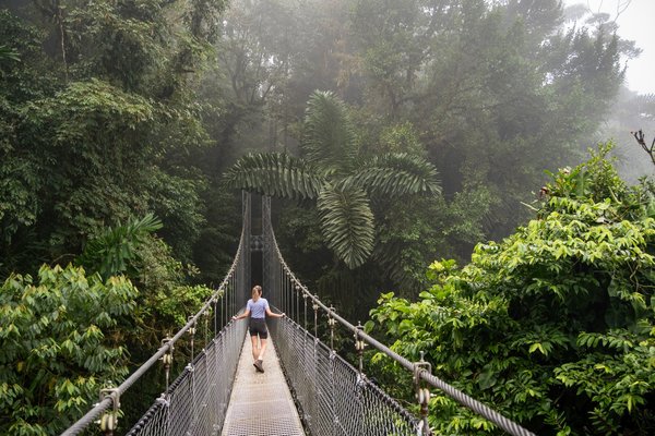 Mujer caminando por un puente colgante rodeado de selva en La Fortuna, Costa Rica, en un día con niebla.