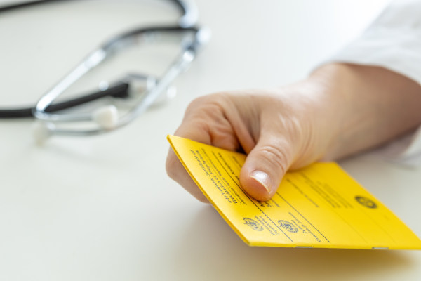 Mano de médico que sostiene certificado de vacunación contra la fiebre amarilla sobre un escritorio al lado de un estetoscopio