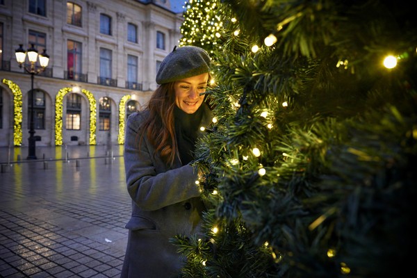 Mujer abrigada disfrutando de las luces de un árbol de Navidad en una calle con ambiente festivo.