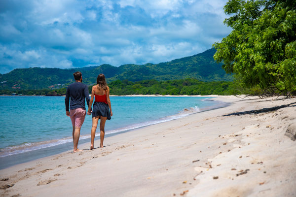 Pareja joven caminando de la mano por una playa de Costa Rica con montañas llenas de vegetación de fondo.