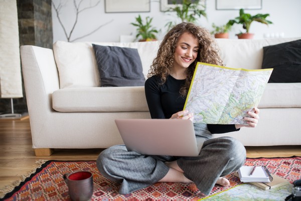 Mujer en el living de su casa mirando un mapa con una laptop en el regazo planificando sus vacaciones con una sonrisa.