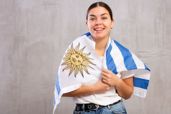Mujer alegre de pie con la bandera de Uruguay colgando en los hombros sobre un fondo liso beige.