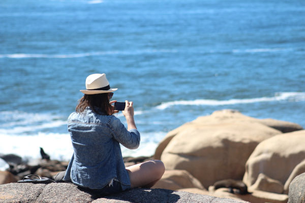 Mujer turista con sombrero y ropa de mezclilla sentada en rocas en Punta del Diablo, Rocha, Uruguay, tomando fotos del mar frente a ella.