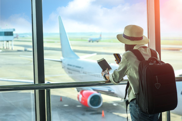 Mujer viajera contra la ventana de una terminal de aeropuerto revisando los vuelos en el teléfono móvil con pasaporte en mano.
