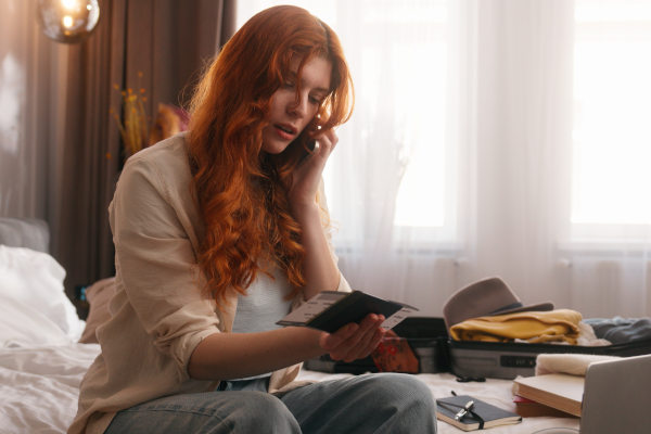 Mujer caucásica mirando documento de viaje mientras habla por teléfono en un dormitorio con maleta abierta de fondo.