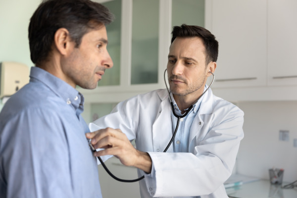 Médico auscultando a paciente masculino de mediana edad por sobre la camisa en el contexto de una consulta médica.
