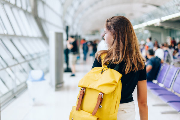 Chica joven de espaldas con mochila amarilla colgada en el hombro esperando viaje internacional en la terminal de un aeropuerto.