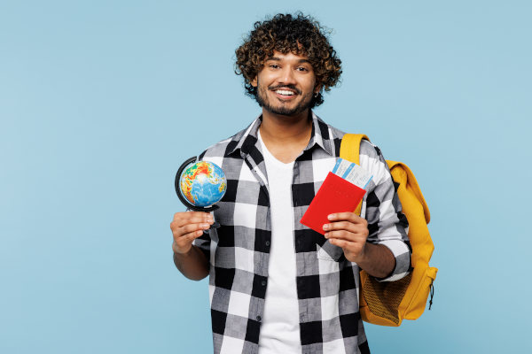 Joven estudiante sonriente sosteniendo globo terráqueo y pasaportes con una mochila colgada al hombro sobre un fondo celeste liso.