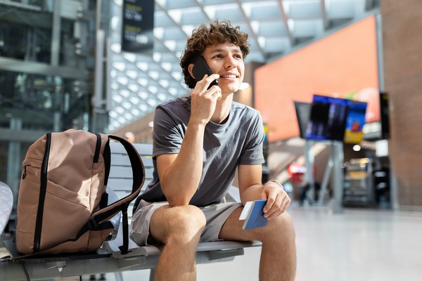 Joven sentado en el aeropuerto hablando por teléfono con el pasaporte en la mano.