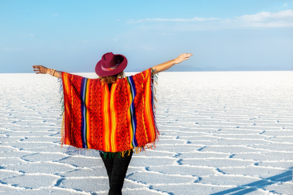Mujer turista con sombrero y poncho extendiendo los brazos en el Salar de Uyuni en un día soleado, Bolivia.