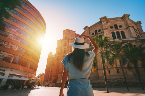 Vista trasera de una joven turista con sombrero blanco de sol caminando por la ciudad de Málaga al atardecer.