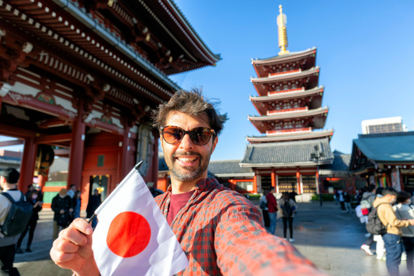 Turista masculino tomándose una selfie con una pequeña bandera de Japón con un templo de fondo en Tokio.