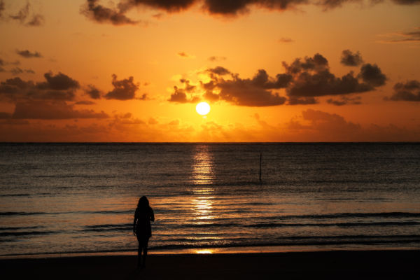 Atardecer en la playa de Maragogi con una turista caminando frente al mar y el sol descendiendo sobre el horizonte.