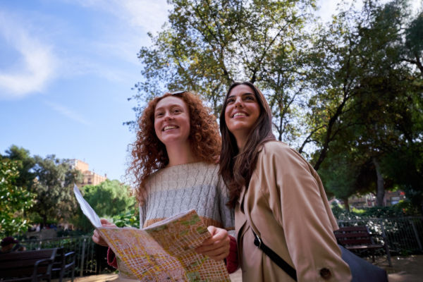 Two young women exploring a European city park with a map in hand, enjoying their vacation under the trees.