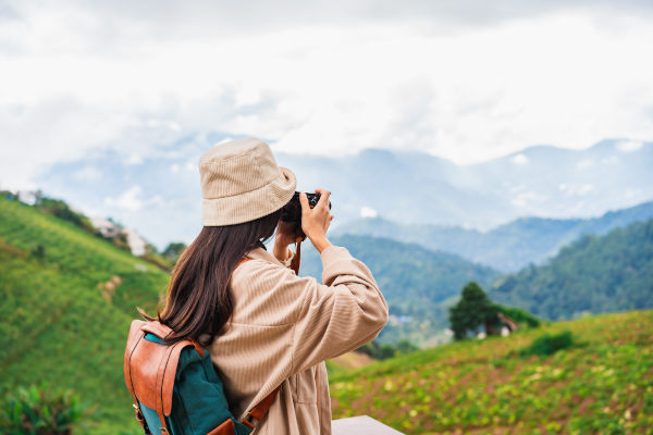 Mujer turista con sombrero y mochila tomando una fotografía con cámara digital de un paisaje montañoso.