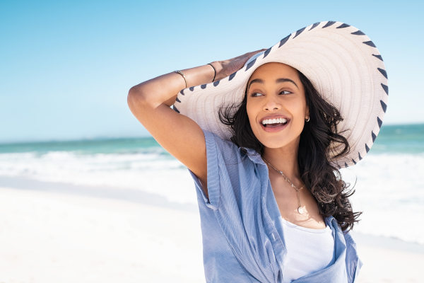 Mujer viajera sonriente sosteniéndose el sombrero en una playa en un día soleado.
