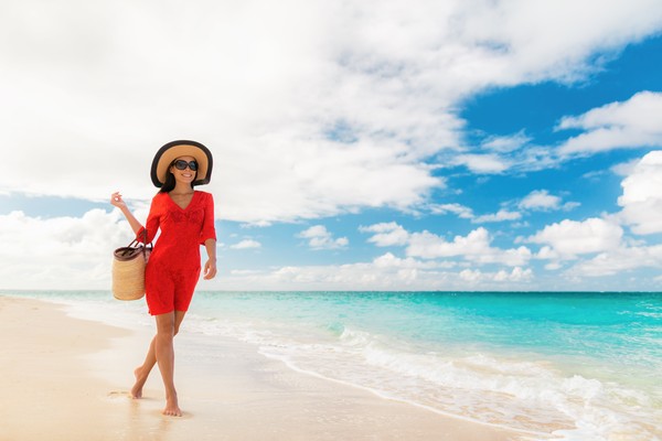 Mujer turista sonriente con vestido de playa, sombrero, lentes de sol y una canasta caminando por una playa caribeña con arena blanca y aguas turquesas