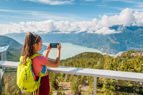 Joven turista con los codos sobre la barandilla de un mirador tomando fotografía con el teléfono de un paisaje natural con montañas y vegetación en Squamish, Canadá.