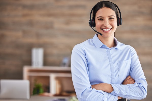 Personal femenino de atención al cliente de pie con los brazos cruzados mientras sonríe a cámara con micrófono y auriculares puestos.