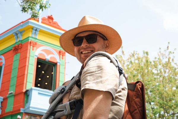 Hombre turista con sombrero y lentes de sol portando una mochila de viaje en la espalda y una mochila portabebés en el pecho.