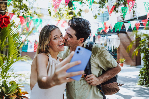 Pareja tomándose una selfie en calle con banderines con los colores de México en un día soleado.
